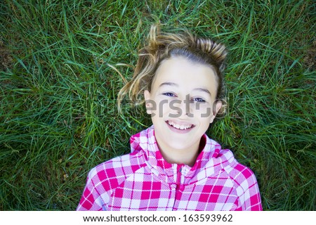 Teenage woman with checked print white hoodie lying back on green grass, laughing and looking at the camera. Candid shot, real people