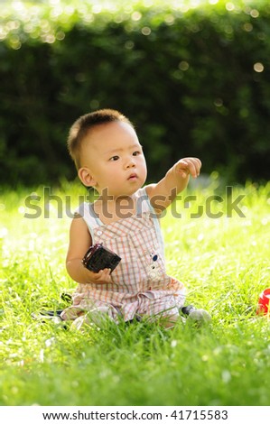 A little Chinese baby boy sitting on the grass with one arm outstretched is pointing his finger