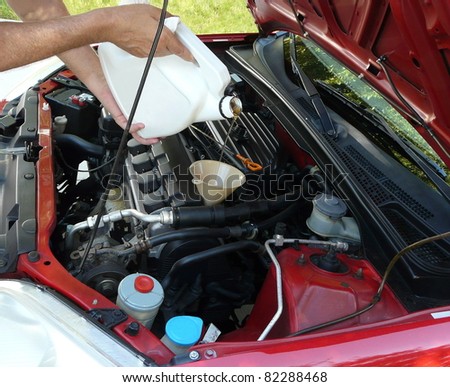 Male adding oil with a funnel from the left side of a red car after a do-it-yourself oil change. Dipstick is nearby.