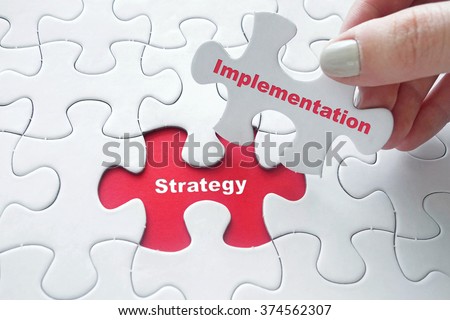 Close up of girl\'s hand placing the last jigsaw puzzle piece with word Strategy Implementation as business concept