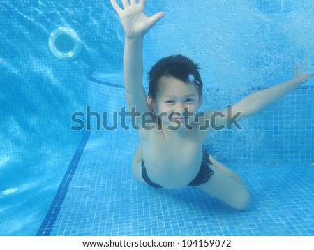 A child boy is smiling underwater in a pool, with his eyes and arms open, without swimming glasses, knees on the bottom of the pool