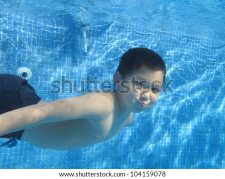 A child boy is swimming and smiling underwater in a pool, without swimming glasses, holding breath, looking at the camera