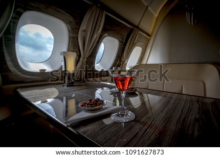 Interior of a business class of a commercial passenger plane, an armchair and a window, a table and a cocktail glass with a drink and almonds. Focus on a glass.