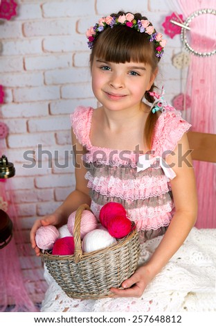 Cute little girl in pink fishnet dress and wreath holding a basket with balls of knitting sitting on a chair in the scenery