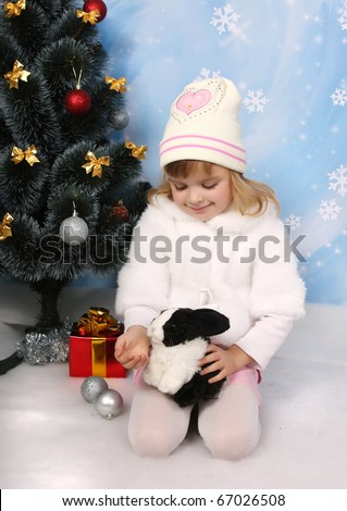 little girl in a white coat and hat with a rabbit around a Christmas tree