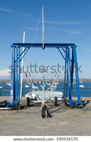A boat trailer positioned under a blue metal hoist in front of a marina with yachts on a sunny day.