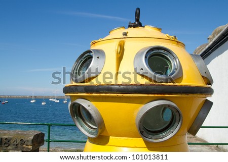 The viewing portals of a vintage diving bell painted yellow with winch attachments the sea and blue sky in the background.