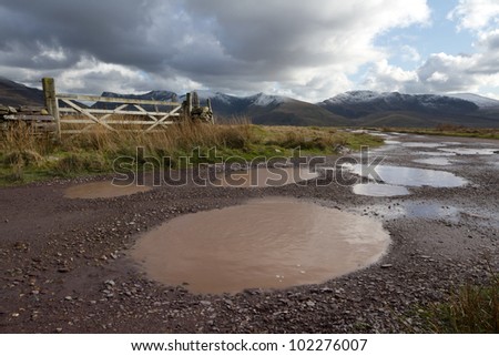 A rough track with water filled potholes cuts through moorland towards a cloudy Nantlle ridge in the Snowdonia national park, Wales, UK.