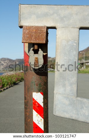 A metal gate, barrier attached to a rusty post with a padlock protected by a steel housing with a tarmac road in the background.