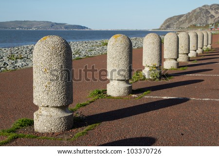 A set of concrete precast parking bollards stretch into the distance with a path, pebble beach and the sea in the distance.