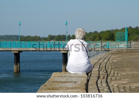 A middle aged, elder woman with short white hair, a white blouse and shoulder strap sits on a wall staring into the distance next to the sea.