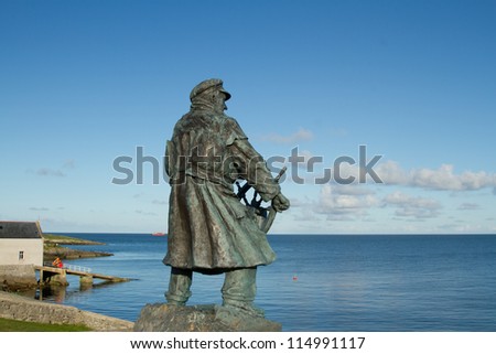 The bronze statue of a sea captain in coat and hat holding a ship\'s wheel looking out to sea with a lifeboat station and ramp in the background.