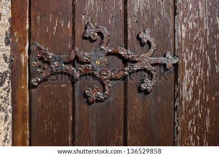 An ornate metal hinge with rust and flaking paint attached to a stained wooden door.