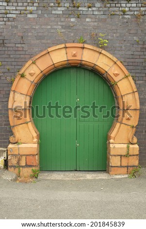 A green painted wooden door with a yellow arch designed with cast brick against an old blue brick wall with plants.