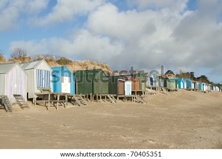 A row of beachhuts, multicoloured, with steps and boardwalks on a beach backed by a sand dune, blue sky and clouds.