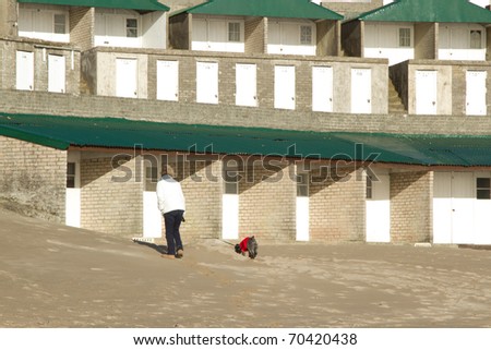 Person, woman, walking a dog in a red jacket on a beach in front of municipal generic beach huts.