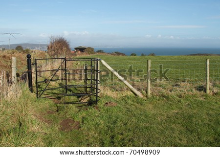 Kissing gate with hedge and fence, public right of way, across agricultural land with green grass, looking towards the sea and sky in the distance.