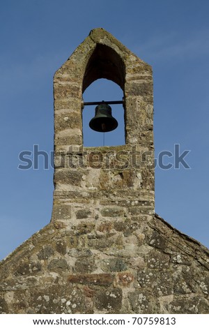 Traditional stone wall and tower houses a bronze bell with bar and rope against a blue sky.
