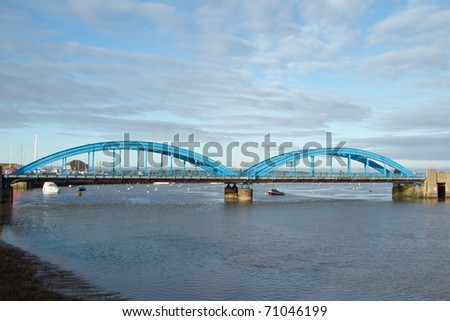 A double span blue painted metal bridge with concrete supports crossing a river with a blue cloudy sky.