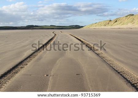Tire tracks going into the distance on a beach with a sand dune and green fields in the distance backed by a blue cloudy sky.