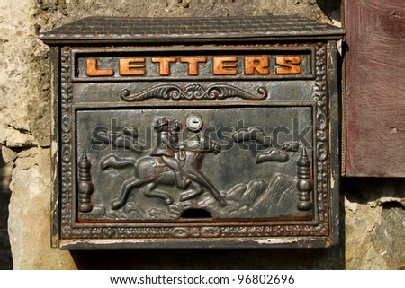 A metal letter box with ornate design painted in black with rust on a stone wall.