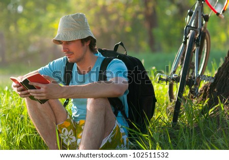 Young traveler with bicycle reading the book while resting in the forest on sunny spring day.