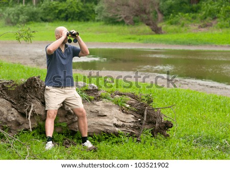 Slim bald man watching the nature through binoculars on cloudy spring day.