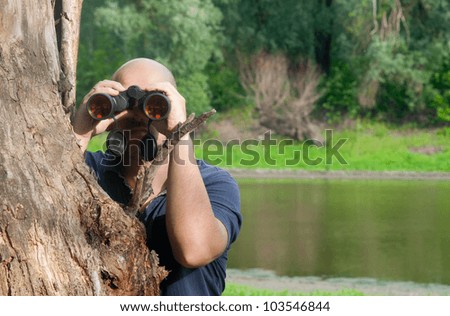 Bald man watching the nature through binoculars while leaning on old tree.