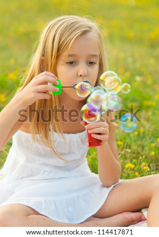 Cute little girl blowing soap bubbles while sitting on the meadow on beautiful spring day.