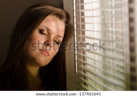 Beautiful sad girl with closed eyes standing beside window with venetian blinds.