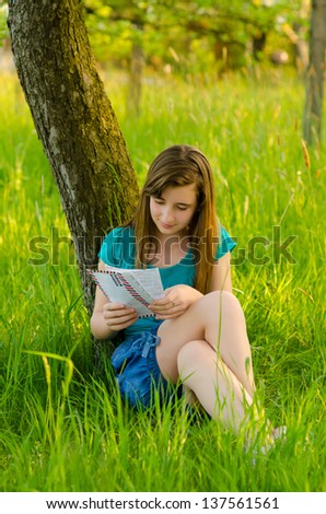 Beautiful teenage girl reading letter while sitting under the tree on sunny spring day.