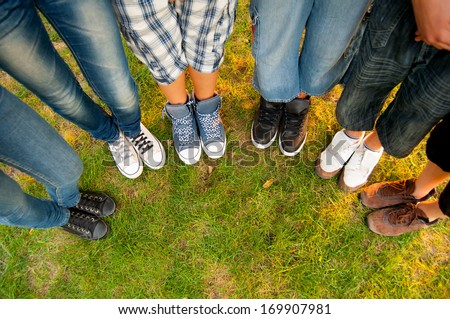 Legs and sneakers of teenage boys and girls standing in half circle on the grass.