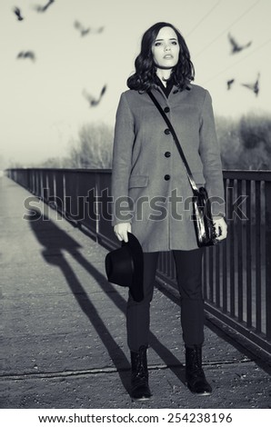Beautiful young fashionable lady standing on bridge at sunrise while birds are flying over her head.