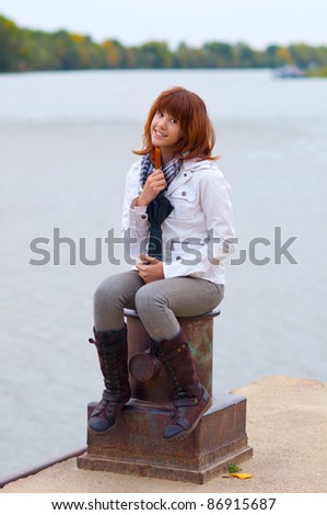 Beautiful smiling redhead teenage girl sitting on the dock column on cloudy autumn day.