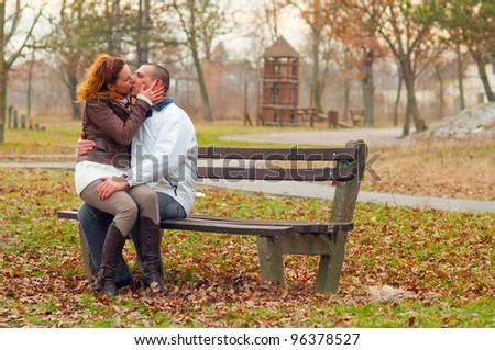 Young couple kissing while sitting on the bench in the park on cloudy autumn day.
