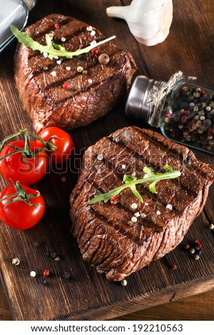 Close up of two juicy thick portions of delicious roasted or grilled beef steak with herbs, peppercorns and spices served on a wooden board with cherry tomatoes in a country kitchen