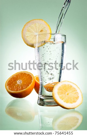 Pouring of mineral water in glass with a lemon and orange on a green background