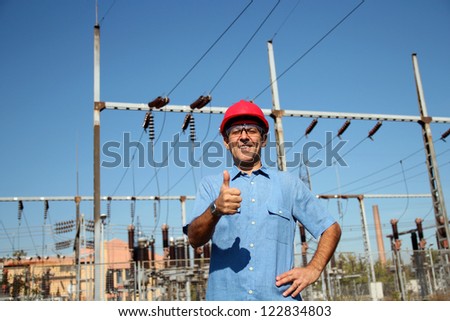 WORKER AT AN ELECTRICAL SUBSTATION. Thumb up given by smiling engineer next to electrical substation. Selective focus.