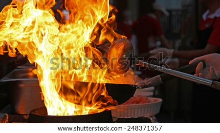 Chef Cooking With Fire In Frying Pan.\
Professional chef in a commercial kitchen cooking flambe style.\
Chef frying food in flaming pan on gas hob in commercial kitchen.