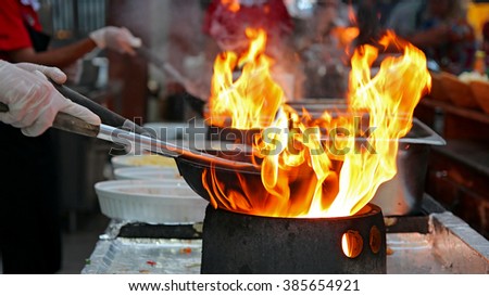 Chef Flambe Cooking. Professional chef in a commercial kitchen cooking flambe style.\
 Chef frying food in flaming pan on gas hob in outdoor kitchen.
