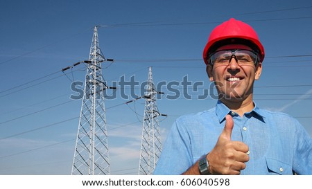 Smiling Worker Near Electrical Transmission Towers. Thumb up given by smiling engineer. A line of electrical transmission towers carrying high voltage. Portrait of an electrical engineer.
