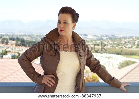 Stylish older caucasian woman in a brown leather jacket, posing casually outdoors, overlooking some houses