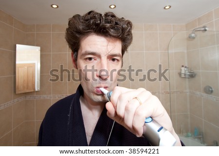 A tired looking man with messy hair brushing his teeth with an electric toothbrush