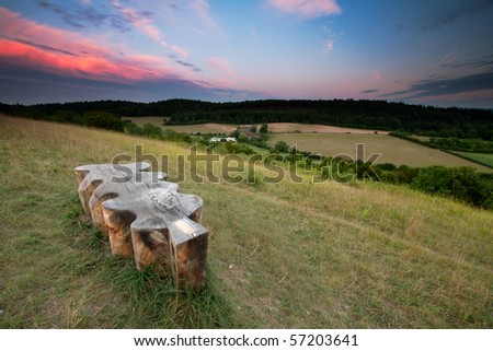 An unusual shaped wooden bench looks over some fields and trees.  The sunset lights up some clouds in the sky.