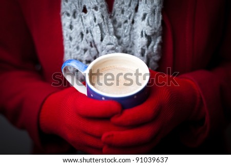 A woman in a red coat and gloves warms her hands on a mug of hot chocolate.  Focus on the drink.