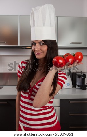 Lovely girl wearing cook cap and holding tomatoes in kitchen.
