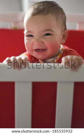Funny little baby with beautiful dark eyes standing in a round white crib and playing just after awakening
