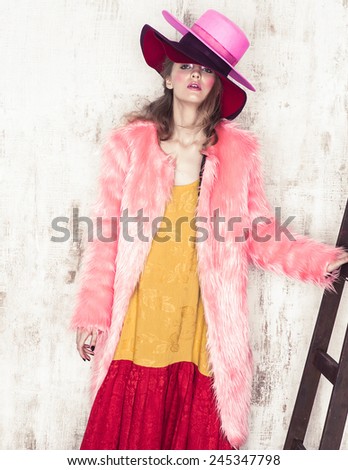 High fashion girl posing near the stair in studio in hat and pink fur coat