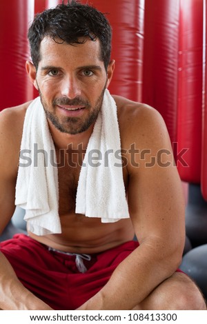 Handsome muscular sports man with a towel on his shoulders smiling in front of red punching bags. Fighter.