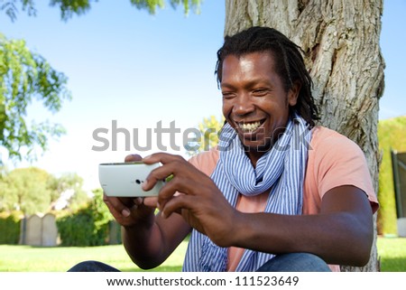 Happy black Jamaican man smiling using an smart phone in the park.